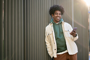 Cool smiling young African American guy holding mobile phone standing at wooden city wall. Happy stylish hipster teenager using cell outdoors, looking at camera with smartphone.