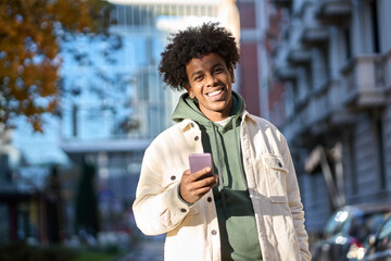 Cool smiling young African American guy holding mobile phone standing with smartphone at big city...