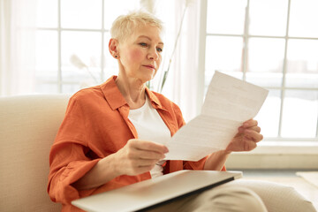 Indoor image of concerned businesswoman of 60s reading paper document sitting on couch in living...