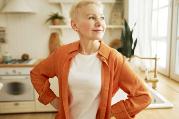 Portrait of pensive mature blonde female of 60 in casual clothes and short haircut standing with...