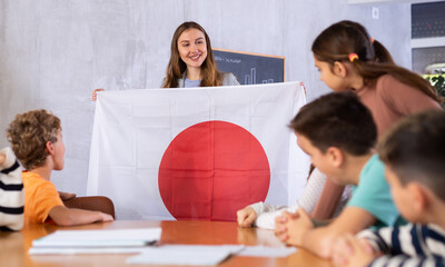 Fototapeta na wymiar Smiling young female teacher explaining culture Japan while showing national flag to schoolchildren preteens in classroom