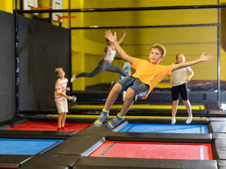 Fototapeta na wymiar Full length of excited casual schoolchild boy having great time while jumping high on colorful trampoline at game club