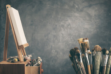 Artistic equipment in a artist studio:  artist canvas on a wooden easel, paint tubes and paint brushes - used artistic paintbrushes for painting with oil or acrylic paints. Copy space for your text. - 573346905