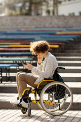 Cute, curly-haired disabled guy in a wheelchair is looking at the phone. A young guy spends his free time outdoors.