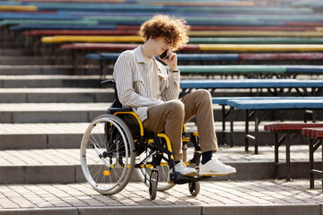 Young disabled man in a wheelchair is talking on the phone. Handsome guy in the park near colorful benches and stairs.