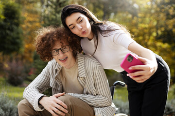 Fototapeta premium Positive, young man in a wheelchair and his girlfriend having fun outdoors while taking a selfie. Young people make funny spades.