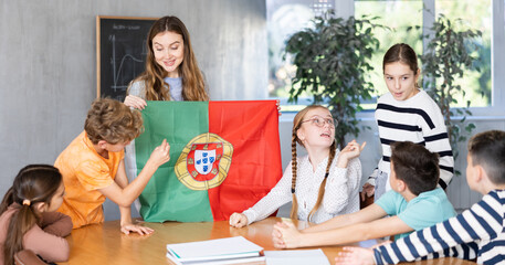 Female teacher, conducting extracurricular lesson in the school class tells schoolchildren the history of Portugal and shows national flag of the country