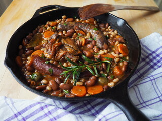French Cassoulet, a meat and beans stew, in a cast iron skillet, on a wooden table with a checkered white and purple dish towel