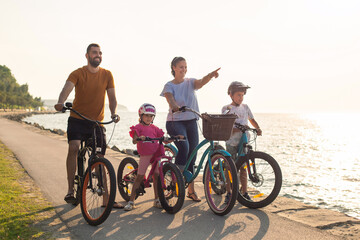 Fototapeta premium Young family, parents with two children, on bikes standing near the sea, looking at view in the direction that the mother pointing