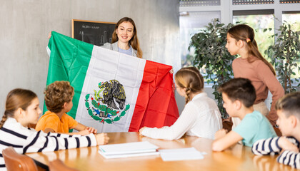 Fototapeta na wymiar Female teacher, conducting extracurricular lesson in the school class tells schoolchildren the history of Mexico and shows national flag of the country