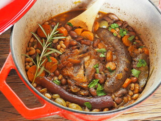 French Cassoulet, a meat and beans stew, in a red cast iron cooking pot, on a rustic wooden table