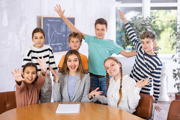 Group photo of joyous teacher and schoolchildren sitting at table and some staying behind in...