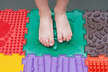 child does leg exercises on orthopedic massage mat at home. Puzzle mats imitating different textures for baby foot massage. Prevention of flat feet and hallux valgus in child. Medicine. Health care.