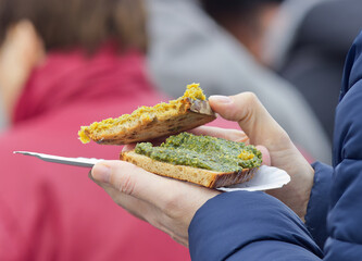 A visitor of Prague farmers market holds a paper tray with a snack, toast with herb pesto.