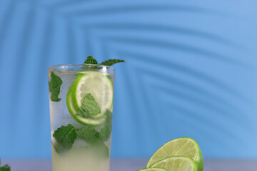 Bright, modern and playful cocktail image of a mojito with green lemon and peppermint with a blue background and a shadow of a palm tree