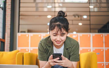 Smiling young asian woman using mobile phone checking social media,app playing game, shopping online, ordering delivery relax while sitting on a couch at cafe with laptop computer