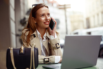 European businesswoman talking phone while working online sitting at outdoors cafe terrace