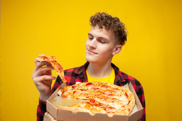 curly cute guy holding pizza box and looking at piece and smiling on yellow background