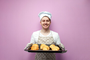 young baker in uniform holding fresh baked croissants and smiling on pink background