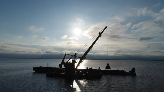 Aerial view. The dredger is working at backlight. Extraction of river sand by a special floating crane. A dredging machine takes sand from the bottom of the river and loads it onto a barge.  