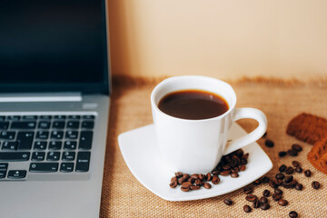 Black coffee in white cup with beans, cookies and laptop on a table. Top view, closeup