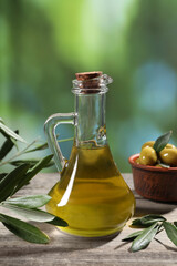 Jug of cooking oil, olives and green leaves on wooden table against blurred background