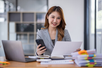 Smiling young Asian business woman executive looking at smartphone using cellphone mobile cell tech, happy ethnic professional female worker working in office typing on cellphone sitting at desk.