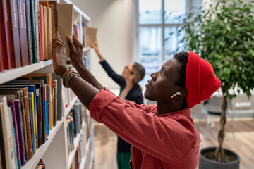 Puzzled serious on finding right educational literature for difficult project student looks through bookshelves in library. African American guy search for book title picks one for lecture class task.