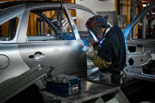 Worker in mask welds metal parts of auto carcass in shop