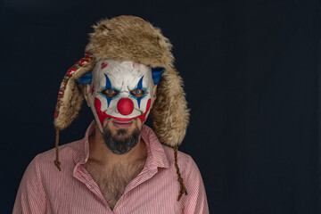A bearded man wearing a creepy clown mask and a hat with ears looks into the camera while standing in a dark room. Scary clown in a red striped shirt in close-up. A maniac clown.
