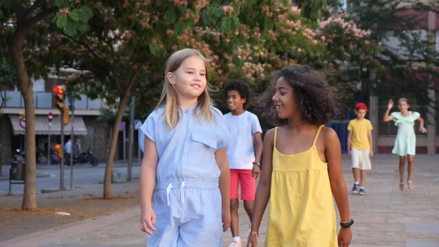 Happy blonde preteen girl walking with her african american girl playmate along city street on summer day 