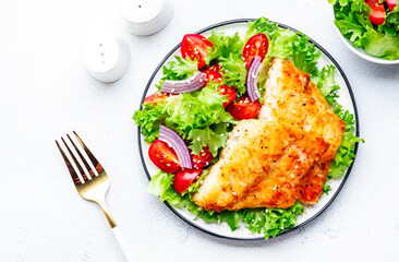 Fried cod fish with salad garnish from lettuce, cherry tomatoes and red onion with sesame seeds, white table background, top view