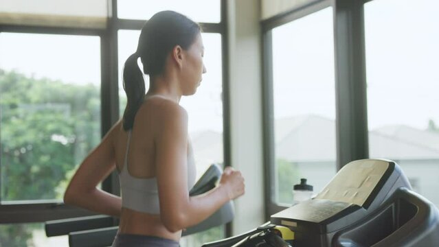 Side view of young beauty Asian female wear sport bra and smartwatch connect with running techno machine start morning routine exercise warming up run in fitness gym at sport club. Girls on treadmill.
