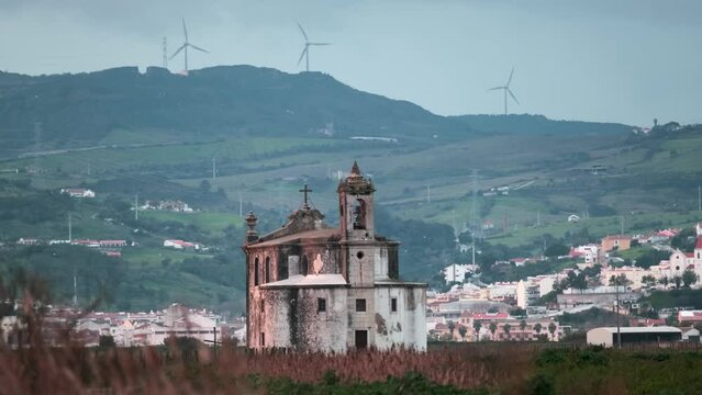Church "Ermida de Nossa Senhora de Alcame" on the background of the city of Lisbon, green mountains and rotating wind turbines with wheat in the foreground at sunset