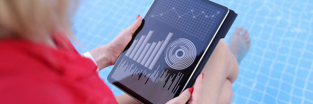 Woman is browsing web statistics using tablet while sitting on edge of swimming pool - Powered by Adobe