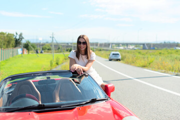 Lifestyle portrait of a carefree beautiful woman in white dress and sunglasses sitting on red cabriolet car and smiling. Road trip enjoying freedom concept 