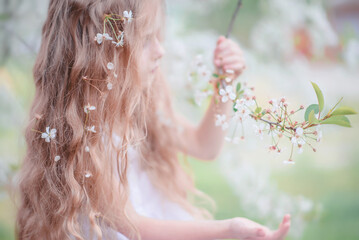 Naklejka premium Spring. A beautiful little girl with long hair and flowers in her hair near a flowering tree. Apple tree in bloom. The Cherry Orchard. Apple orchard. The baby is holding a twig. Portrait. tender baby
