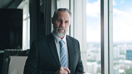 Smilling face of a senior businessman wearing a suit stading near a large glass window looking at...