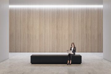 Front view on blank wooden wall background with space for poster presentation in spacious abstract hall with businesswoman using laptop and sitting on black bench on concrete floor, mock up - Powered by Adobe