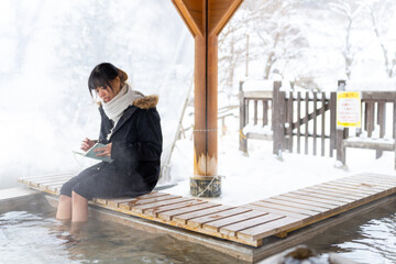 Asian woman soaking feet in hot water and writing journey memories in diary book. Attractive girl resting at wooden seat pavilion covered in snow during travel village in Japan on winter vacation. - Powered by Adobe