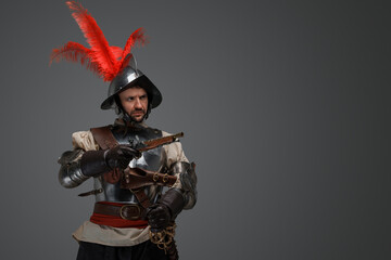 Portrait of antique proud musketeer dressed in steel plate armor holding pistol.