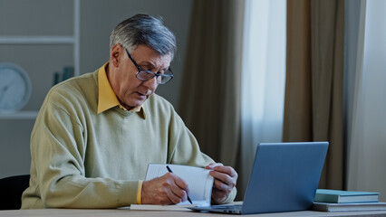 Caucasian grandpa 60s male businessman old senior mature man grandfather writing information in diary notebook letter make handwriting notes working at home office looking laptop software application