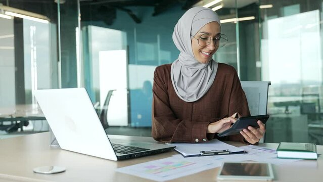 Positive muslim accountant clerk in hijab work with papers at desk in modern glass office Concentrated bookkeeper count financial report on calculator with documents and laptop computer at workplace