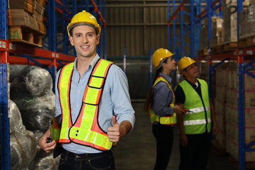 Group of warehouse workers with hardhats and reflective jackets using tablet, walkie talkie radio and cardboard while showing thumb up and smiling in retail warehouse logistics, distribution center