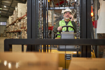 Front view of bearded male worker driving forklift truck in warehouse and talking to portable radio © Seventyfour