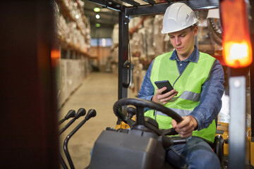 Portrait of young worker driving forklift truck in warehouse and using smartphone