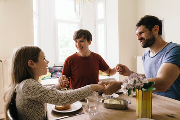 Happy family holding hands sitting at dining table in home