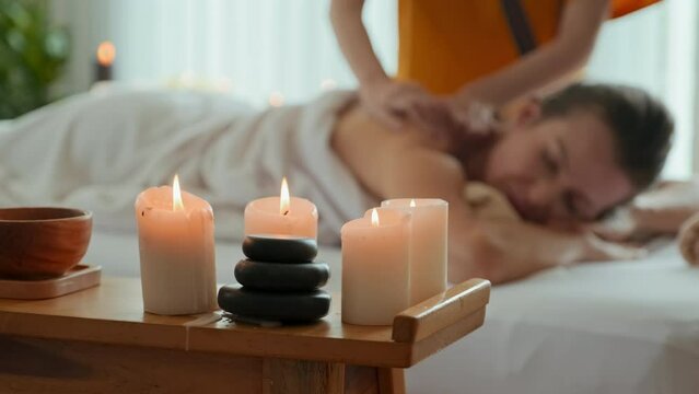 Young beautiful Caucasian woman relaxing during back massage in luxury spa room with burning aromatic candles in focused foreground