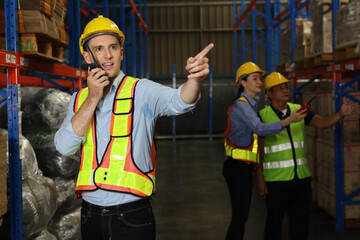 Group of warehouse workers with hardhats and reflective jackets using tablet, walkie talkie radio and cardboard while pointing something in retail warehouse logistics, distribution center