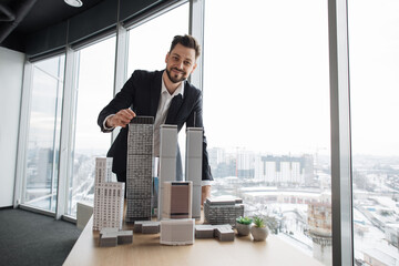 Confident businessman real estate agents in black stylish formal suit standing while touching 3d model skyscraper of city architecture design at office with panoramic city view.